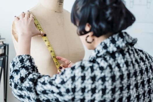 Back view of crop anonymous female tailor in checkered wear with flexible ruler measuring chest of dummy in workshop