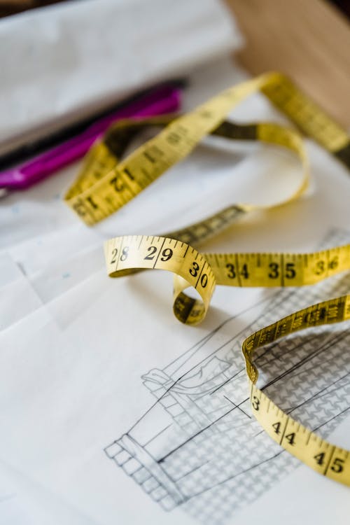 From above of long yellow measuring tape placed on table with papers and draft of attire in light modern workshop