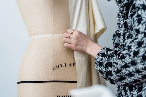 Crop unrecognizable female seamstress in warm clothes standing near professional mannequin with unfinished attire in modern atelier on white background