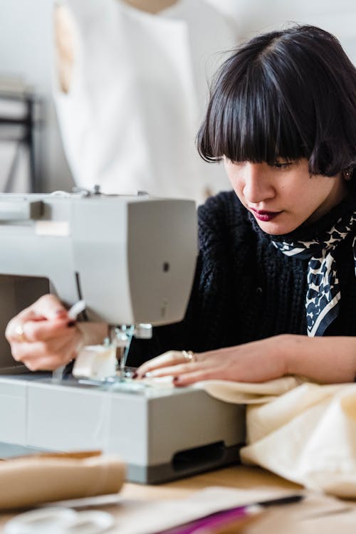 Concentrated Asian female tailor with black short hair sewing textile on machine while working in light modern workshop with mannequin