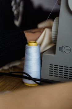 Crop anonymous female dressmaker sewing textile on sewing machine at wooden table with spool of white thread in modern atelier