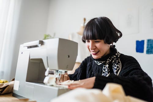 Free Happy young ethnic female dressmaker with stylish haircut smiling while sewing new garment on machine during work in light atelier Stock Photo