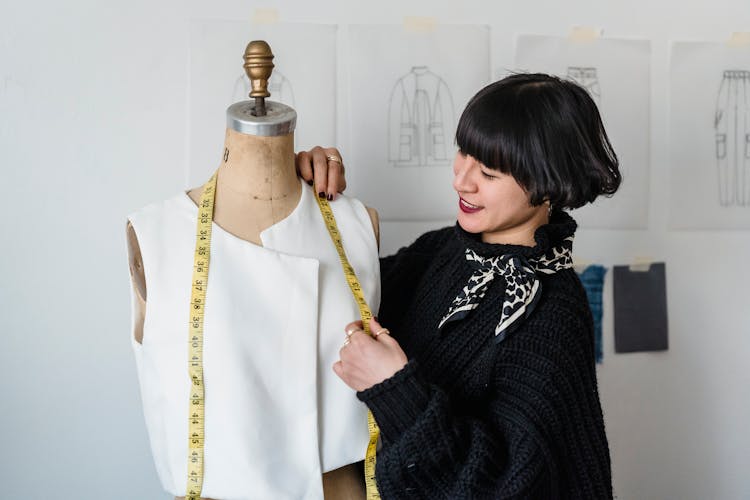 Smiling Young Asian Female Dressmaker Taking Measurements From Mannequin