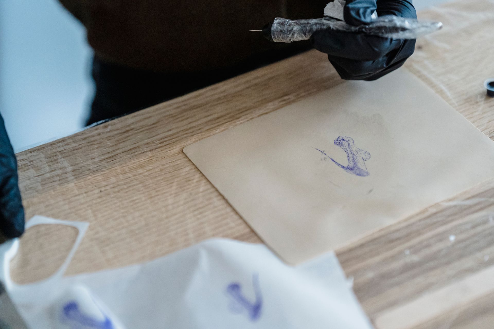 A gloved person holds a wrapped tattoo machine above a sheet with a small blue stencil design on a wooden table.