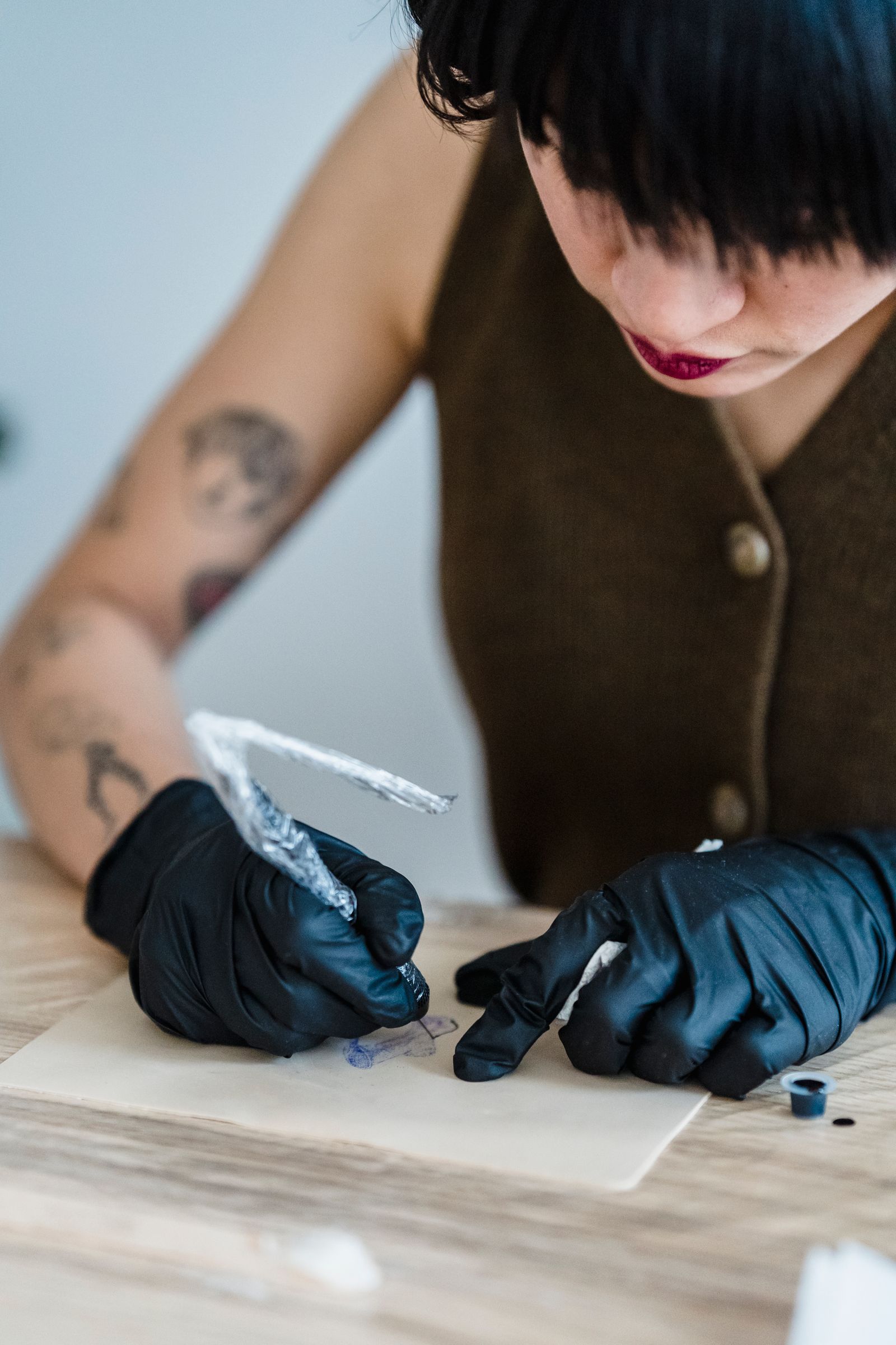 A tattoo artist wearing black gloves uses a tattooing tool on a design laid out on a work surface.