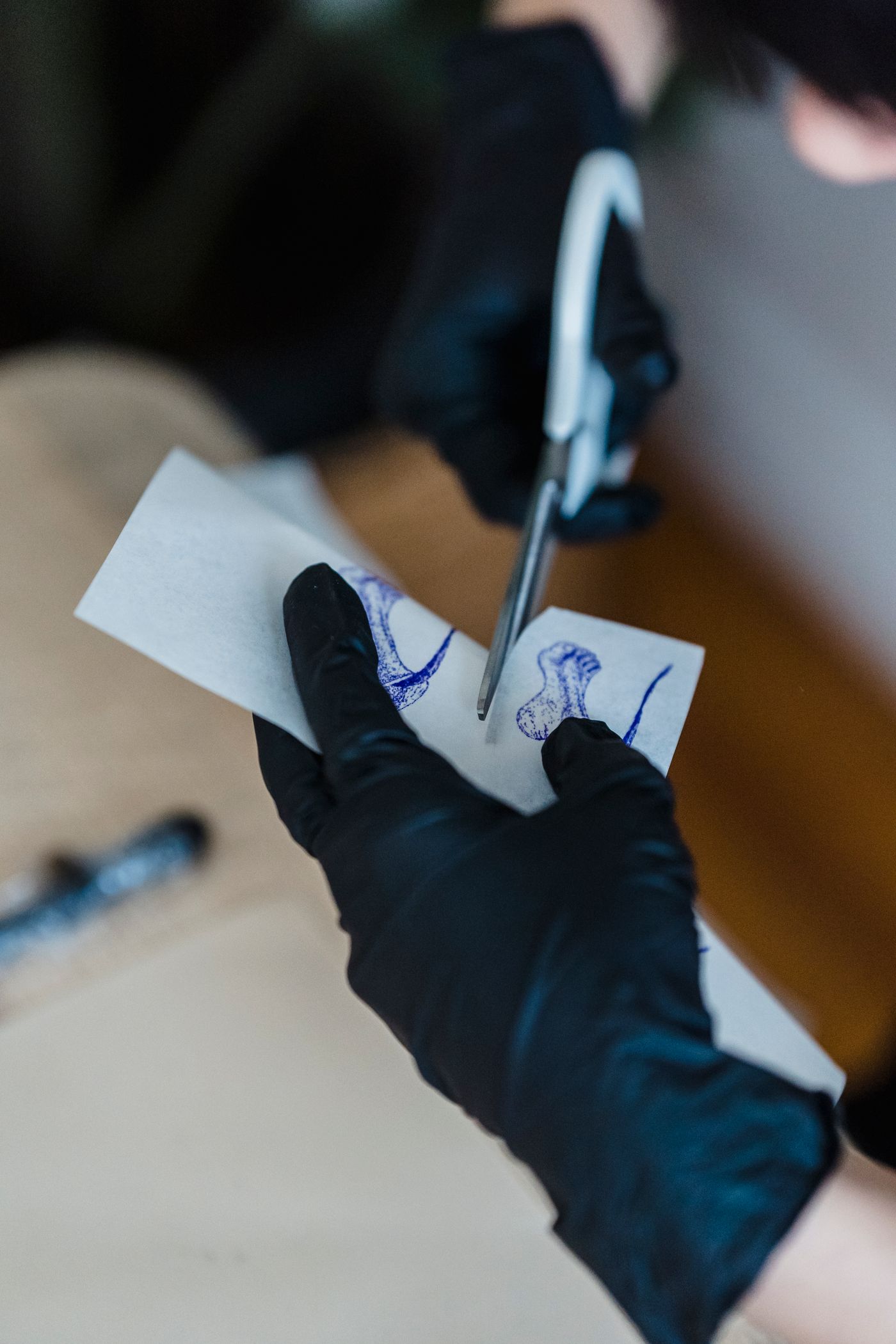 A gloved tattoo artist holds scissors over printed tattoo design paper on a work surface.