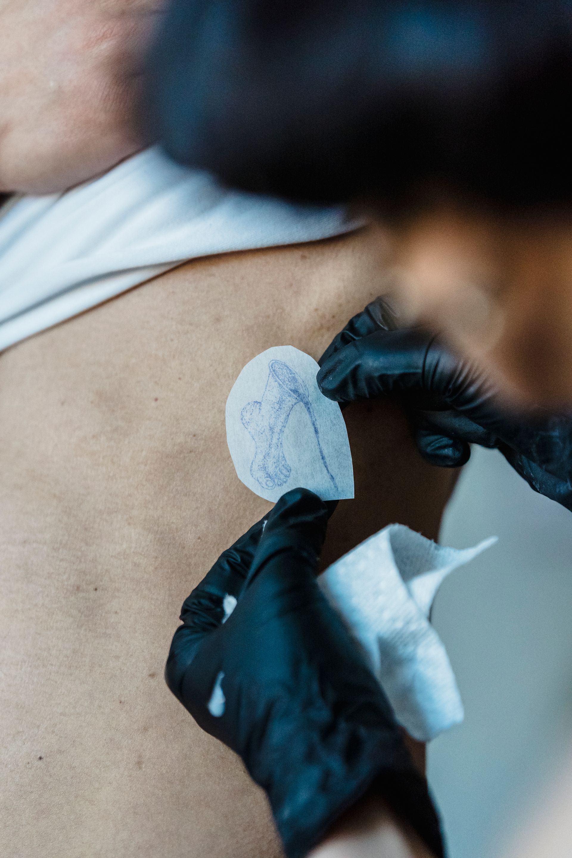 Gloved hands hold a small blue tattoo stencil or design transfer against a person’s skin while preparing the placement.
