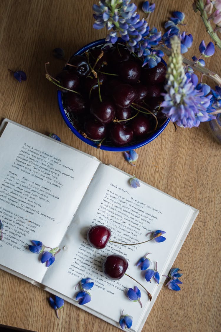 Opened Book Placed On Table With Cherries And Blue Flowers