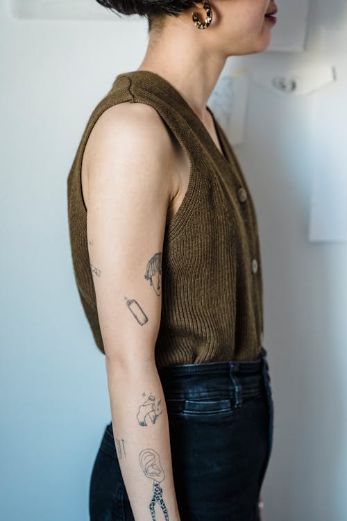 Side view of crop unrecognizable lady with short dark hair and creative tattoos on arms in trendy outfit standing against white wall