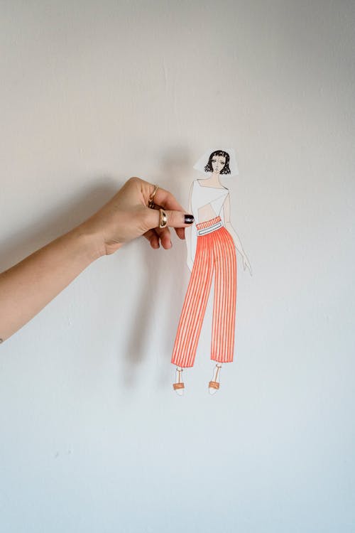 Crop anonymous female dressmaker holding cut out fashion illustration against white wall while creating new collection in light atelier