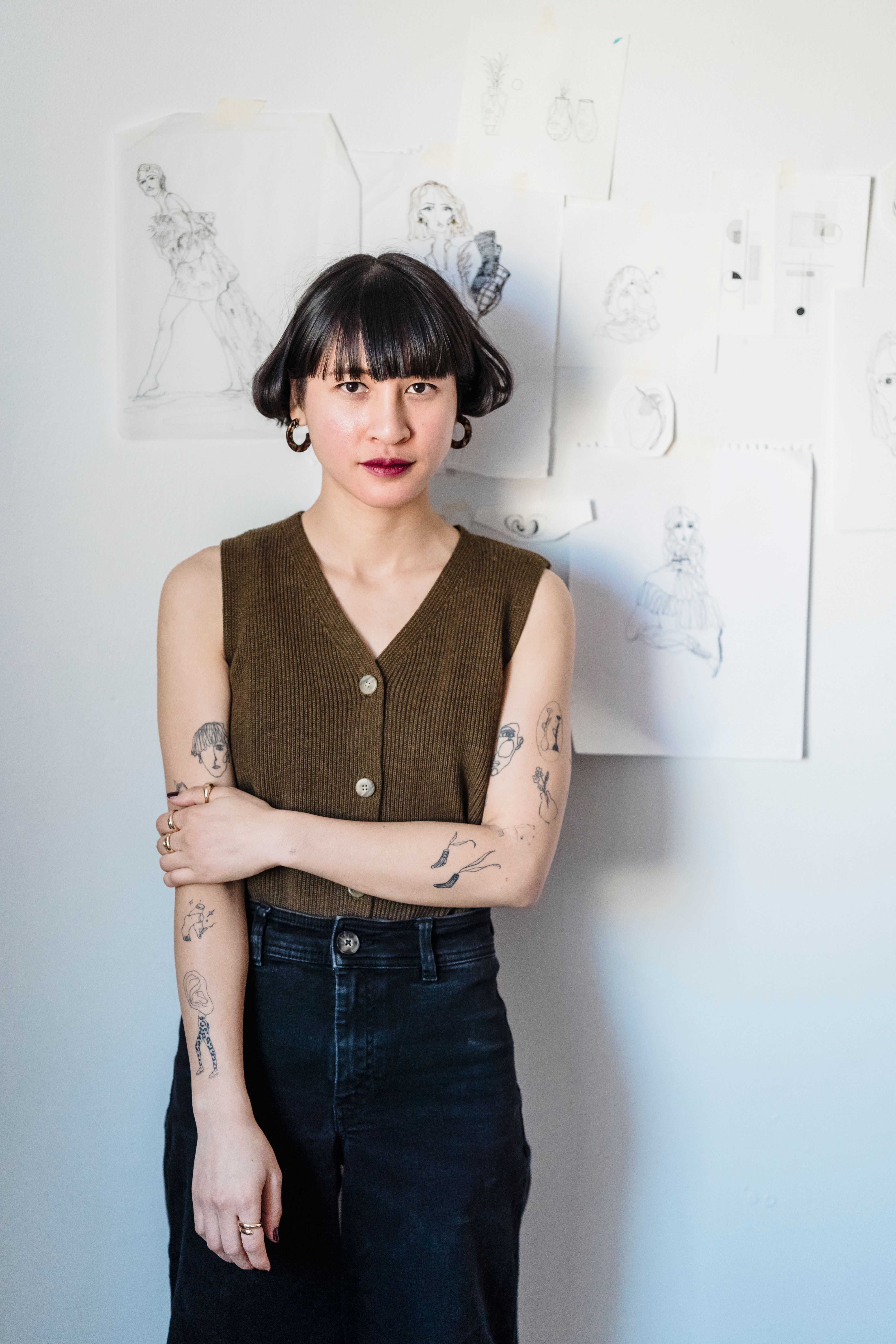 Calm trendy young ethnic female millennial with short dark hair and tattoos on arms standing near wall with papers with fashion illustrations and looking at camera