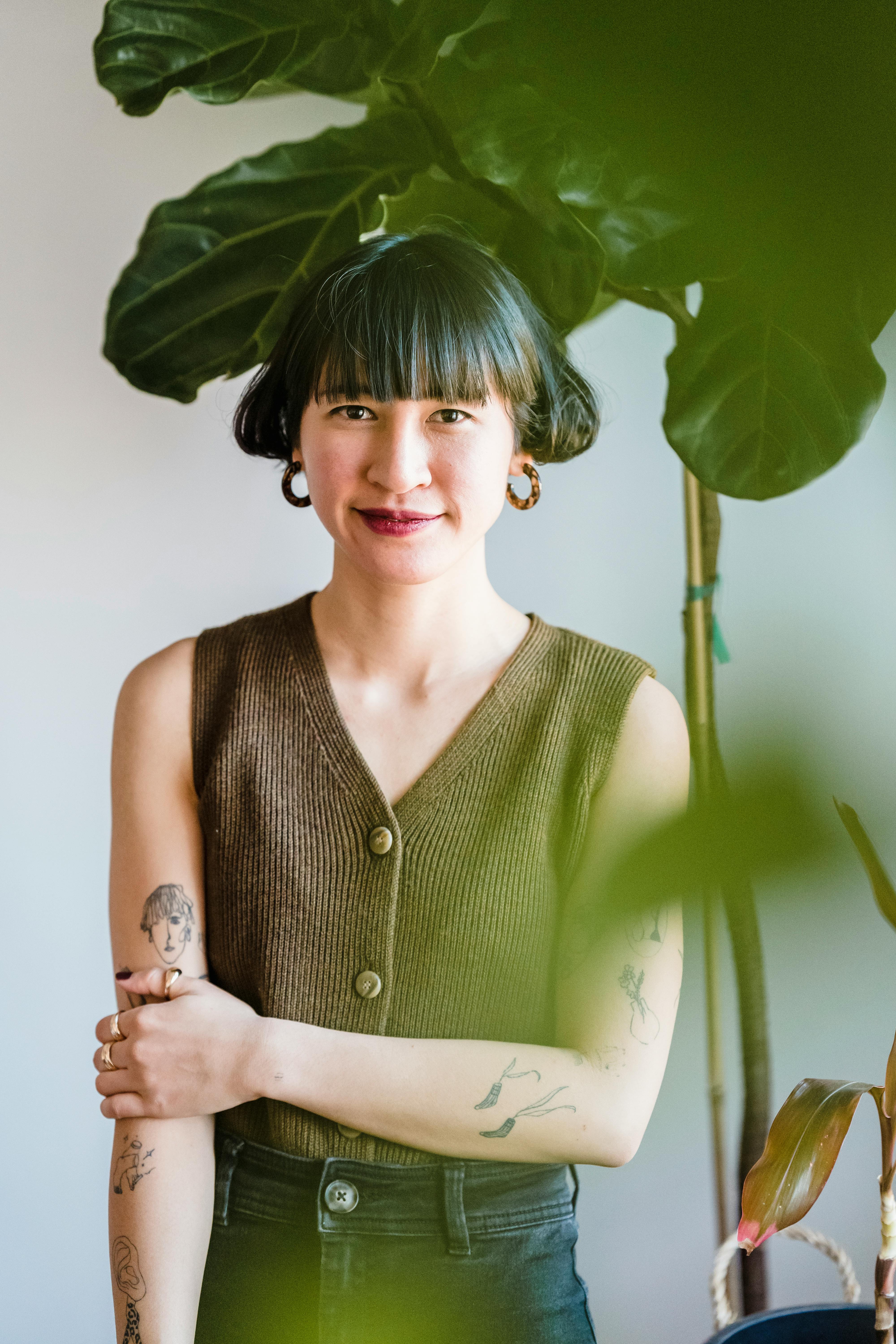 Positive young Asian female millennial with trendy hairstyle and tattoos on arms smiling and looking at camera while standing near big potted plant in light room