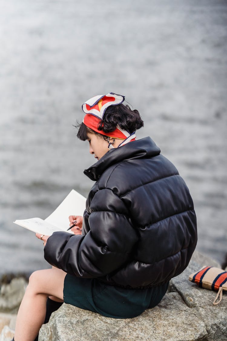 Young Woman Sitting Near River And Drawing In Notebook In Daytime