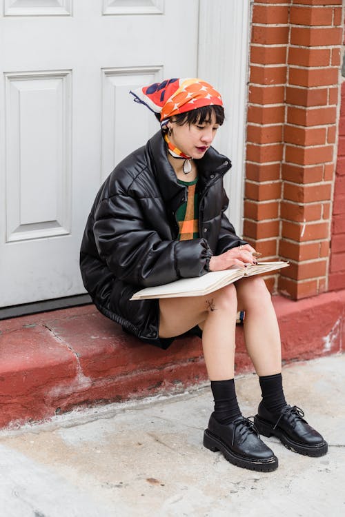 Full body of young trendy female pondering and working with planner white resting on step of brick building