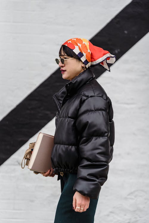 Side view of female in stylish round shaped sunglasses and black warm jacket looking away near white wall with black stripe