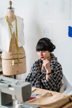 Pensive young Asian female designer sitting at workplace with sewing machine against mannequin in light studio in daytime