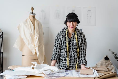 Positive Asian female dressmaker with measuring tape on neck standing at wooden table with fabrics during work in atelier in daytime