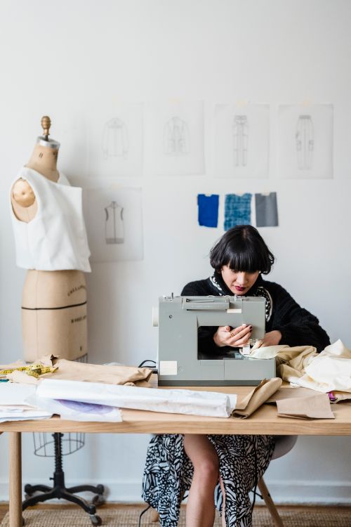 Free Concentrated Asian female dressmaker sitting at table and sewing on sewing machine against wooden mannequin in light atelier Stock Photo
