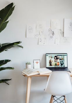 Laptop and opened book placed on wooden table near light wall with paintings in apartment in daytime