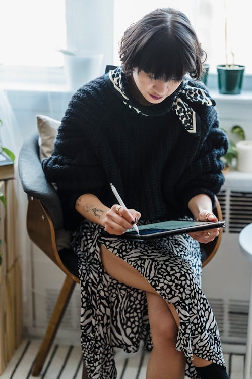 Concentrated young female illustrator in stylish outfit sitting on chair and drawing on graphic tablet in light room against window in daytime