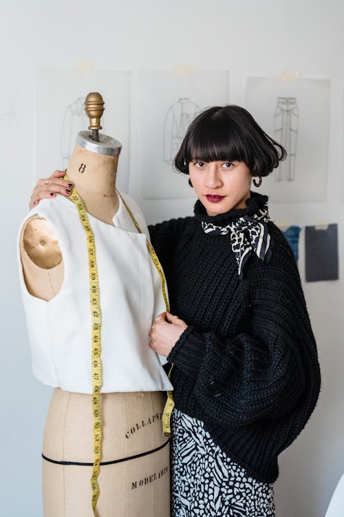Confident young Asian female designer with short hair in stylish outfit standing near mannequin with measuring tape and looking at camera in atelier in daytime