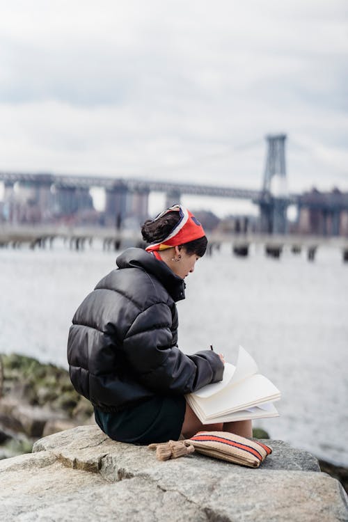 Side view of unrecognizable lady in warm clothes sitting on rock and taking notes in notepad near river and bridge under cloudy sky in daytime