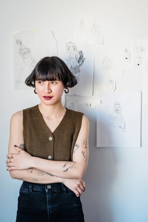 Woman with tattoos near wall with papers with sketches