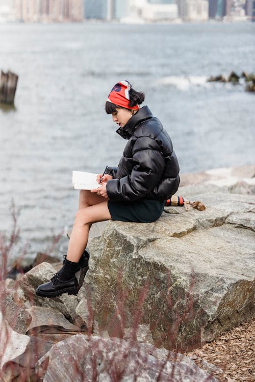 Side view of focused female student in warm casual clothes taking notes in planner while sitting on stone on river embankment