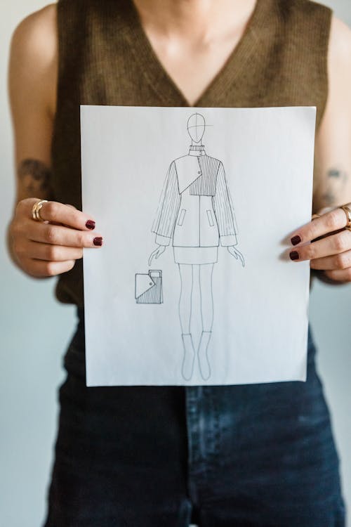 Crop unrecognizable female fashion designer wearing casual clothes demonstrating paper with drawing of new creative model of jacket and purse