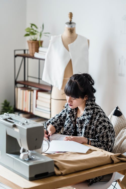 Free Side view of concentrated female tailor in stylish clothes sitting at table with sewing machine and making sketches in light modern atelier Stock Photo