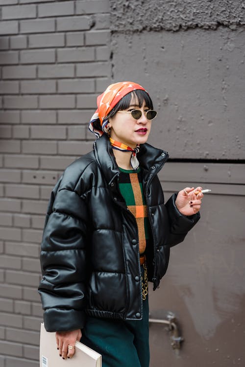 Young female in trendy outfit and sunglasses smoking cigarette while walking on street near brick wall and looking forward