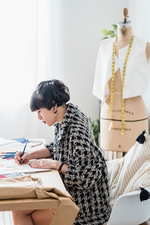 Side view of focused female dressmaker in trendy outfit drawing sketch while sitting at table in light workshop