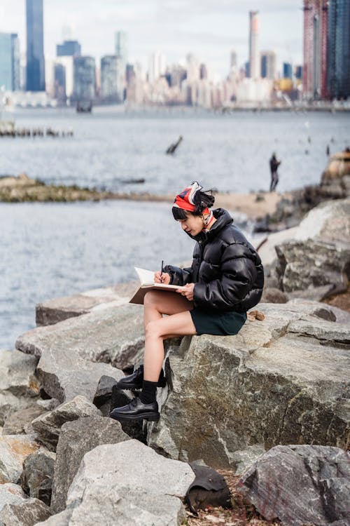 Side view full body of young female in trendy outfit sitting on rocky coast of river while taking notes in daylight