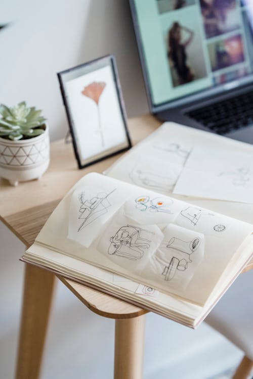 From above of opened notebook with different sketches placed on table with potted plant and photo frame near laptop in light room