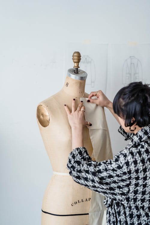 Free Side view of crop unrecognizable female in trendy outfit trying on white fabric on mannequin in light workshop Stock Photo