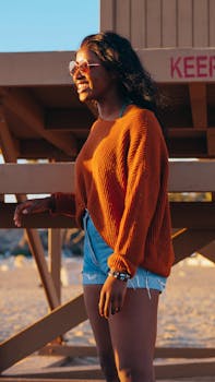 Happy woman wearing sunglasses and sweater by the beach, enjoying the sunlight.