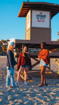 Three women socializing near a lifeguard tower on a sandy beach during golden hour.