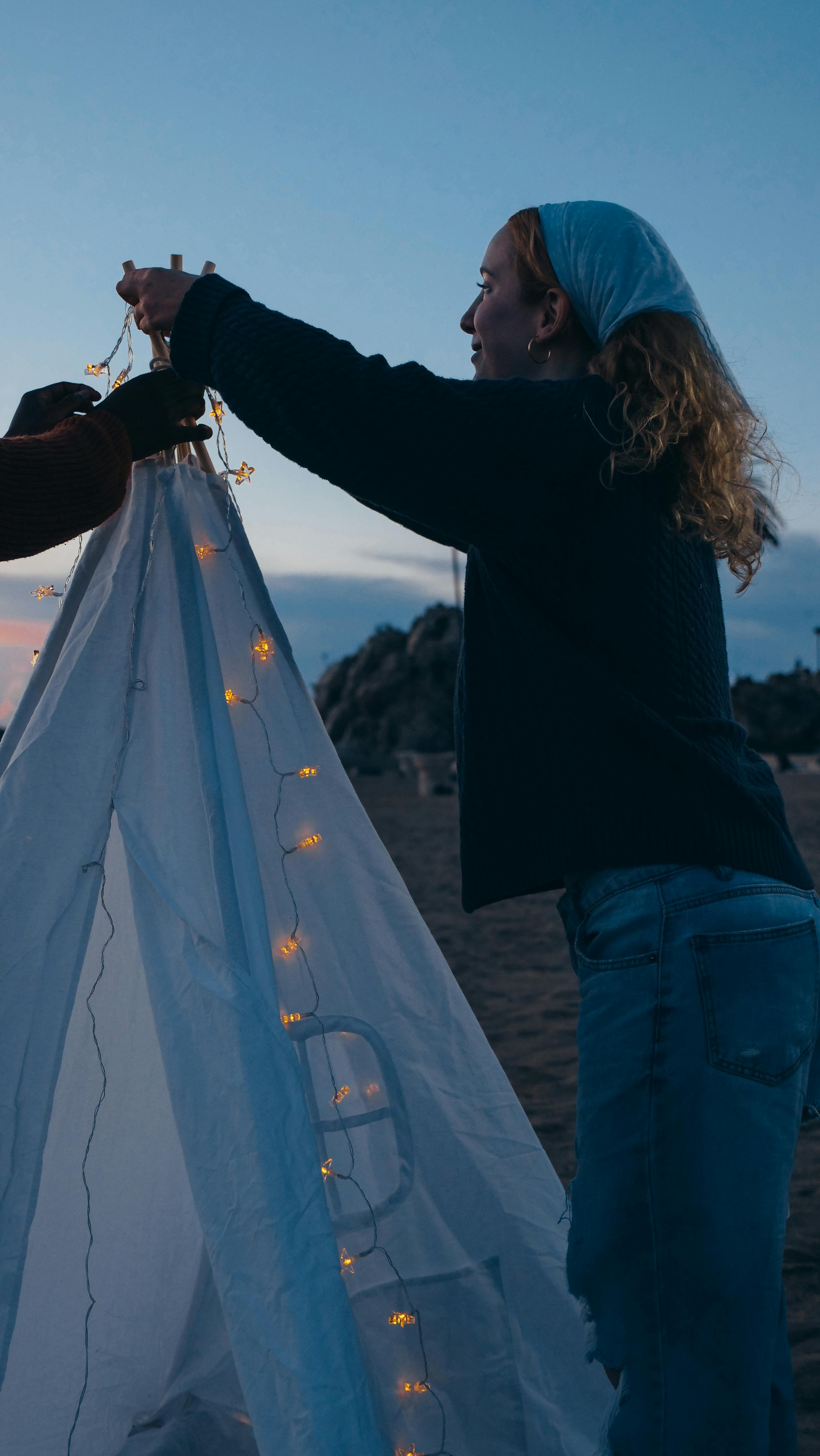 A woman decorates a teepee with string lights on a beach at dusk, creating a cozy atmosphere.