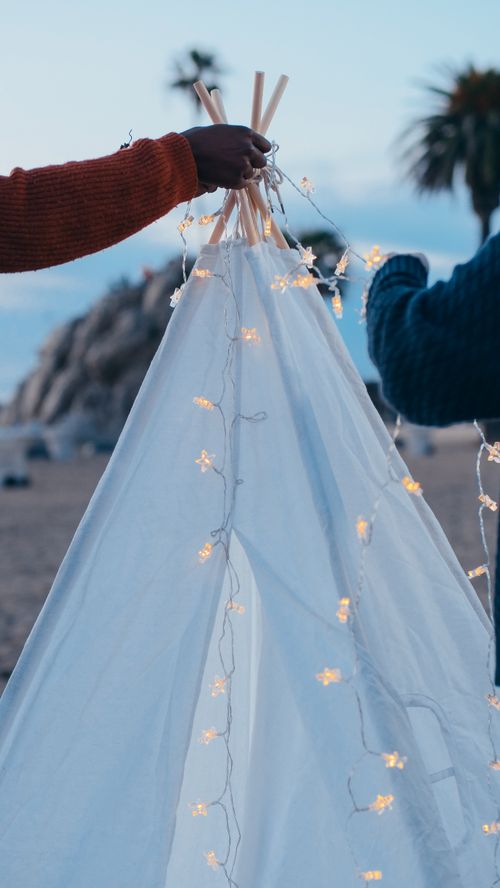 Free A cozy white teepee tent is decorated with string lights on a serene beach setting at dusk. Stock Photo