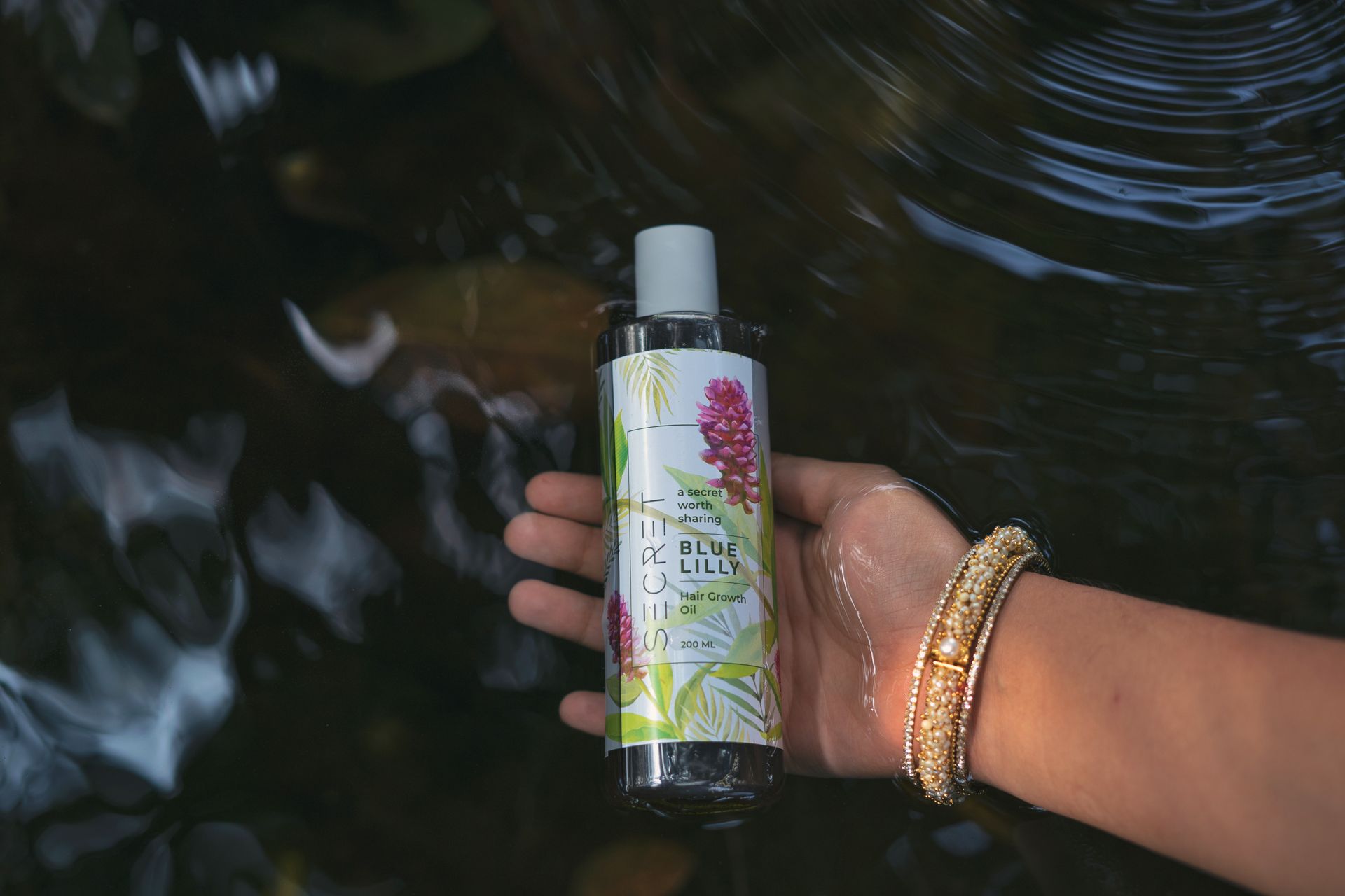 Holding a Bottle of Blue Lilly Hair Growth Oil on the Surface of the Pond