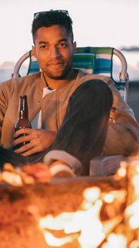 A man enjoying a cold beer while sitting by a campfire at sunset by the shore.