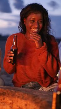 A woman happily holding a beer bottle at a beach bonfire during the evening, conveying relaxation and enjoyment.