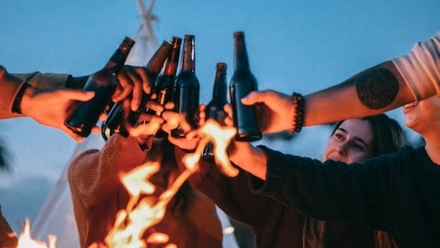 A group of friends enjoying a bonfire, clinking beer bottles in a joyful sunset gathering.