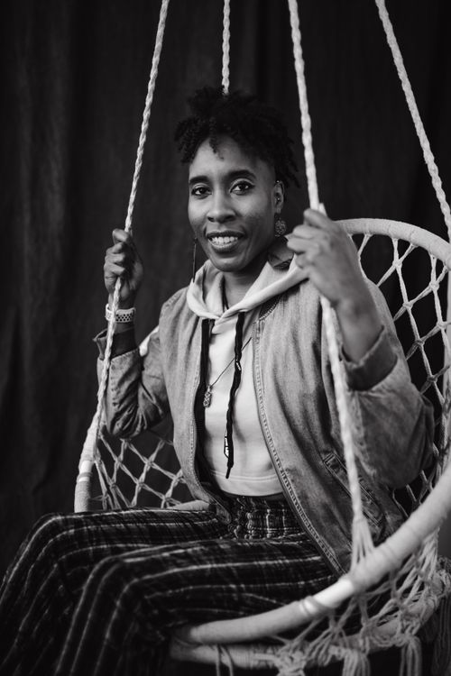 Free Black and white portrait of a woman with afro hair smiling while sitting on a hanging chair indoors. Stock Photo