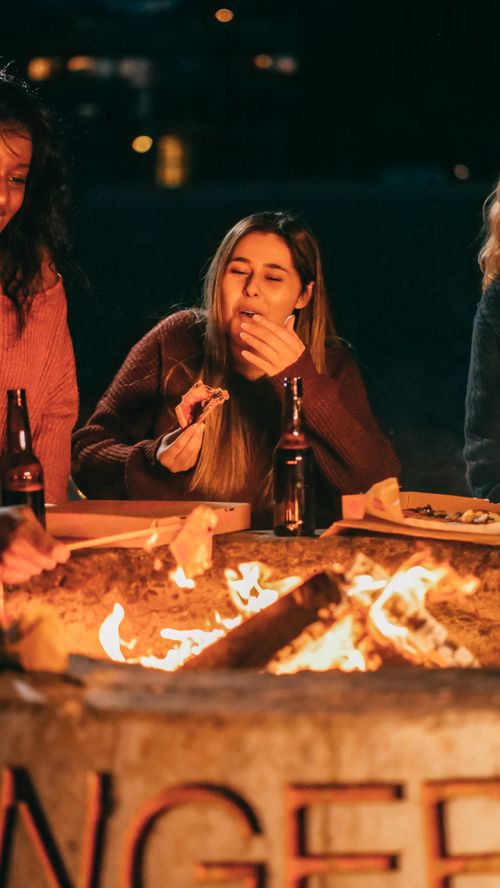 Free Group of friends enjoying pizza and beer by a cozy bonfire at night on a beach. Stock Photo