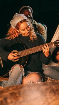 A joyful young woman plays guitar at a campfire, surrounded by friends, enjoying the evening.