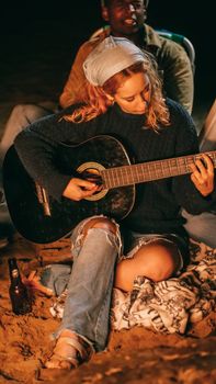 Young woman plays guitar around a beach bonfire with friends under the night sky.