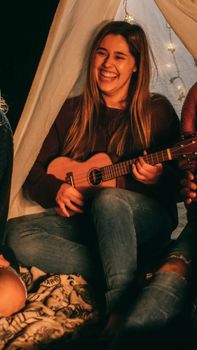 A happy woman playing ukulele inside a tent during a cozy night gathering.