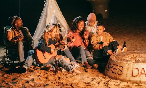 Free Group of friends enjoying music and socializing by a bonfire at night on the beach. Stock Photo