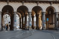 Unrecognizable citizens on asphalt roadway with tram rails under aged masonry construction with arches and colonnade in city